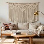 A bright living room featuring a large white macrame wall hanging with heart design above a beige linen sofa, rattan coffee table with books, potted fiddle leaf fig plant, terracotta pots, and gold arc lamp.