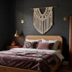 Bedroom interior with gray walls, a rattan headboard on an oak bed frame draped in purple velvet bedding and a colorful woven throw, macrame wall hanging, carved wood mirror, brass wall lamp, wooden nightstand, potted fiddle leaf fig plant, and dark curtains over a window.