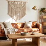 A boho living room with tan leather sofa, white knit pillows and throw, wooden coffee table, macrame wall hanging above limestone fireplace, plants, ladder shelf with baskets, and patterned rug on wood floor.