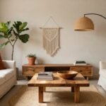 Bright living room featuring a white linen sofa with orange pillow, low wooden coffee table topped with books and wooden bowl on round seagrass rug, fiddle leaf fig plant in terracotta pot beside built-in wooden bookshelf, black arc floor lamp, and white macrame wall hanging.