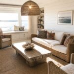 A bright living room featuring a white slipcovered seagrass sofa, two woven armchairs, a rustic wood coffee table on a jute rug, and built-in bookshelves beside a large window.