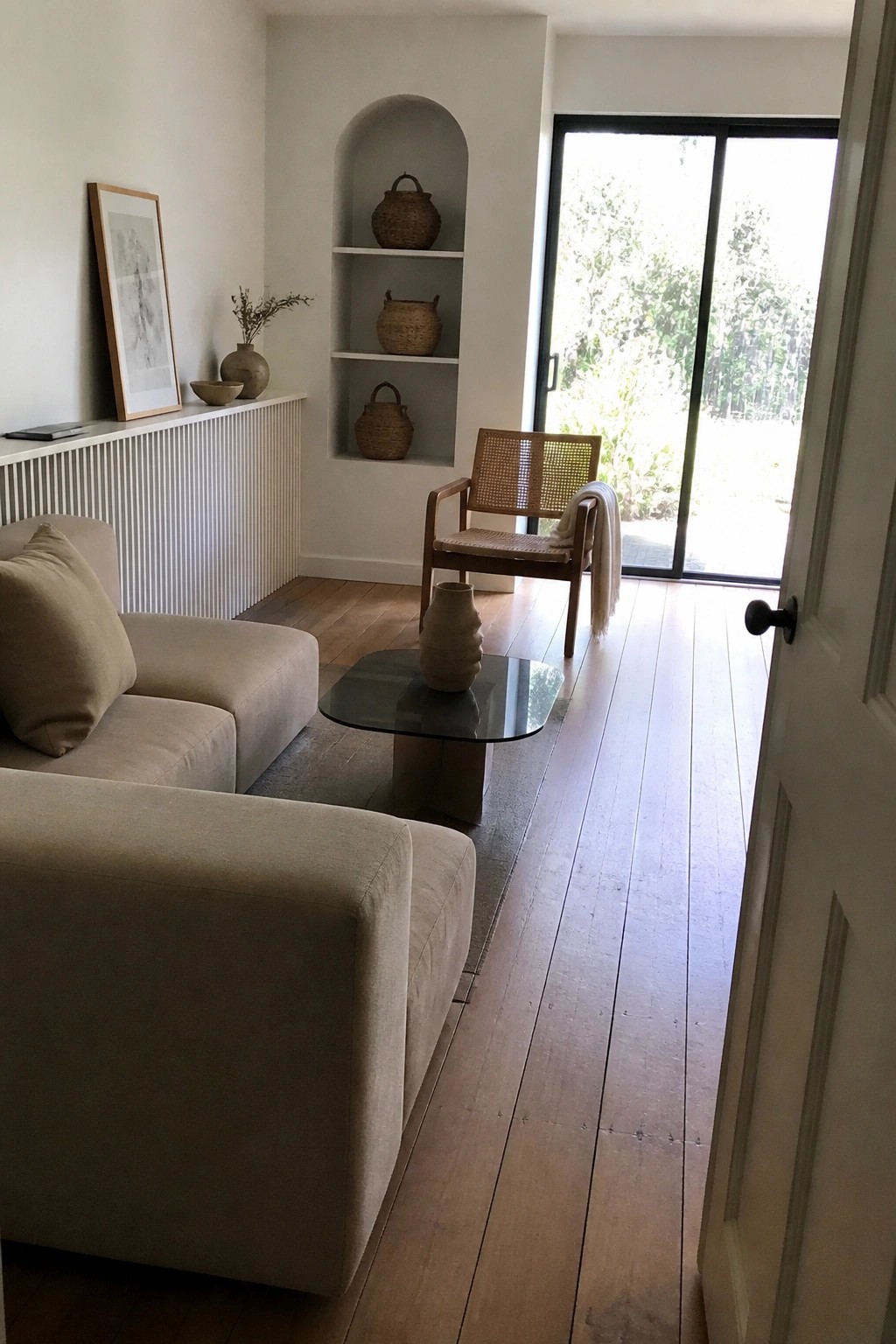 A bright minimalist living room featuring a beige sectional sofa, glass coffee table, cane-back wooden chair, and built-in arched wall shelves holding woven baskets and pottery.