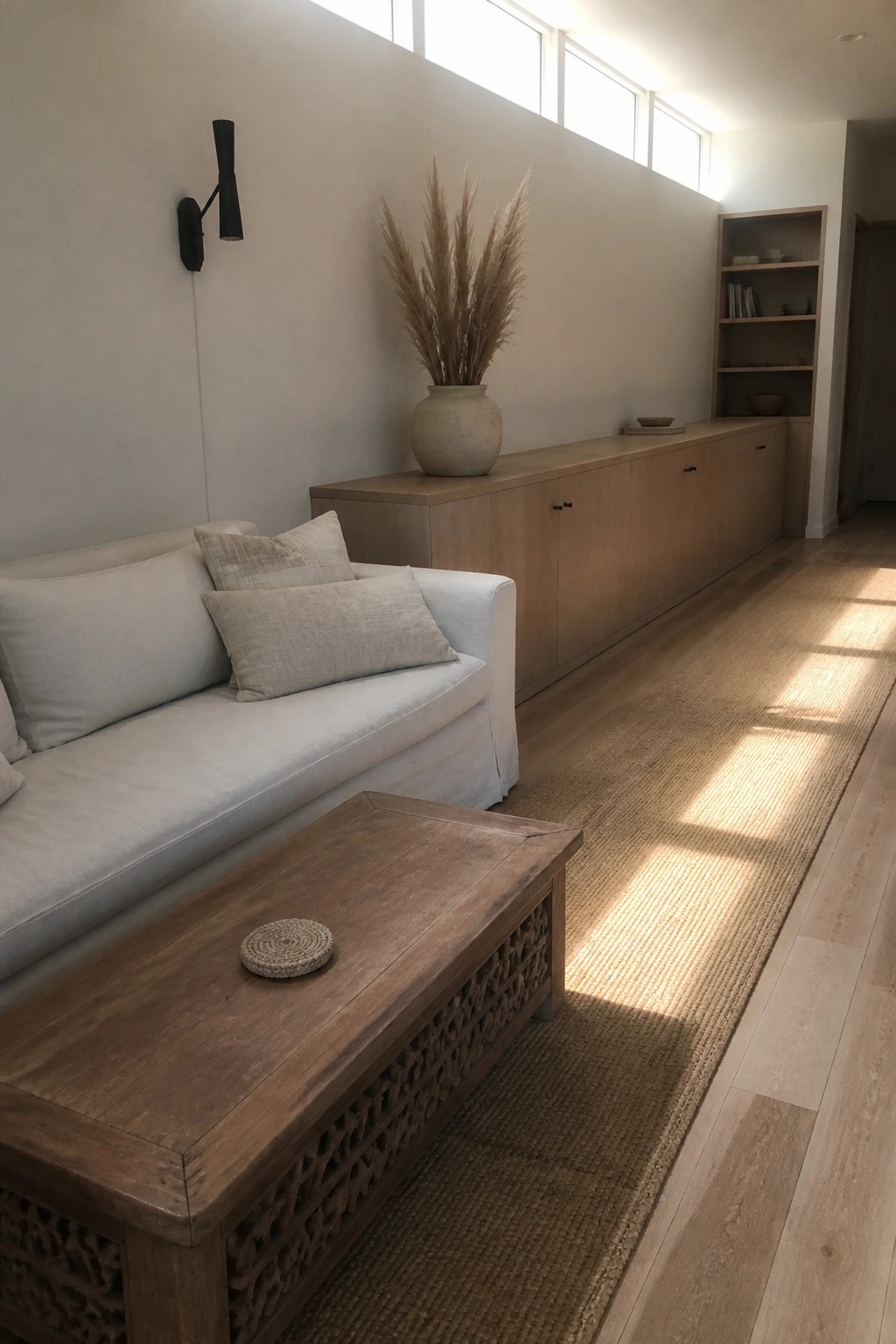 A minimalist living room featuring a white sofa, carved wooden coffee table, long built-in light wood cabinet, and large vase with dried grass under tall windows.