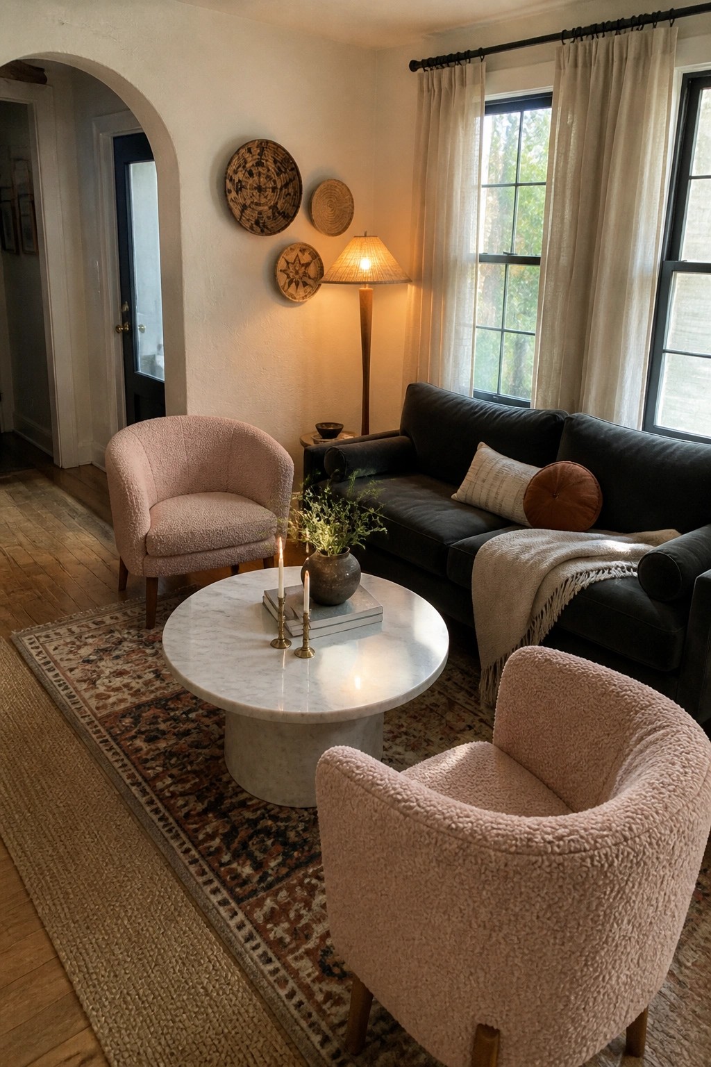 Living room featuring two pink bouclé accent chairs, a dark sofa, round marble coffee table with candles, and woven wall decor on a white wall.