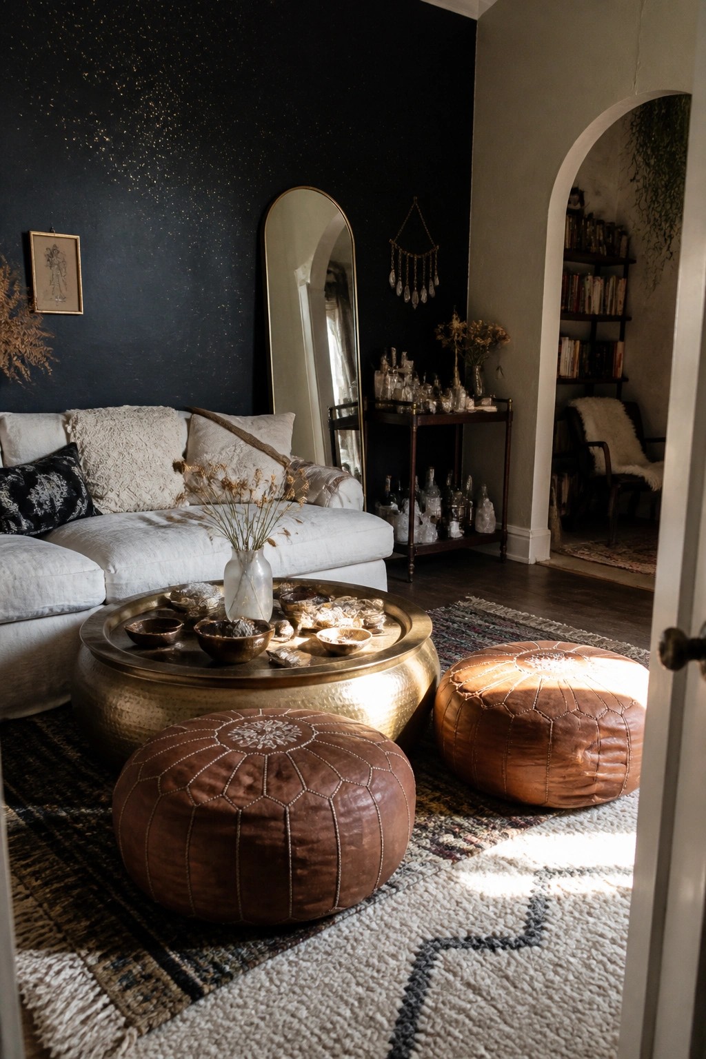 A living room featuring a dark navy wall with gold speckles, a white sofa, two brown leather poufs, a round brass coffee table, and an arched mirror.