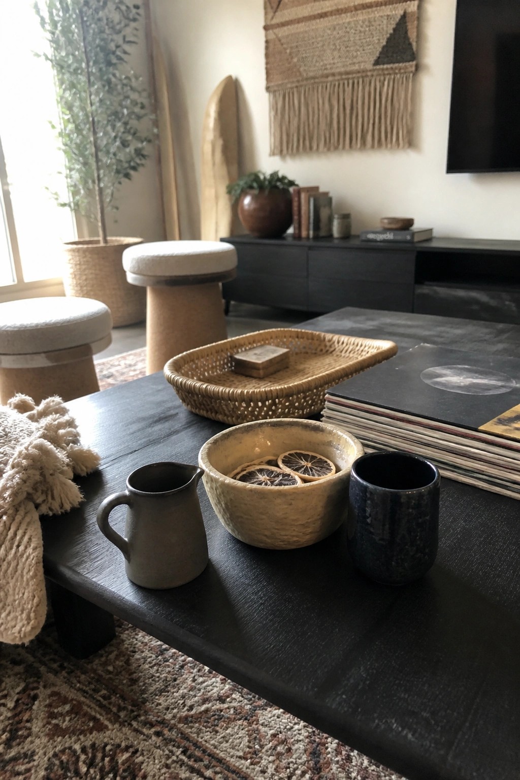 A dark wood coffee table holds a small gray ceramic pitcher, a beige ceramic bowl with dried orange slices, and a dark blue ceramic cup, with a woven tray and stacked records nearby.