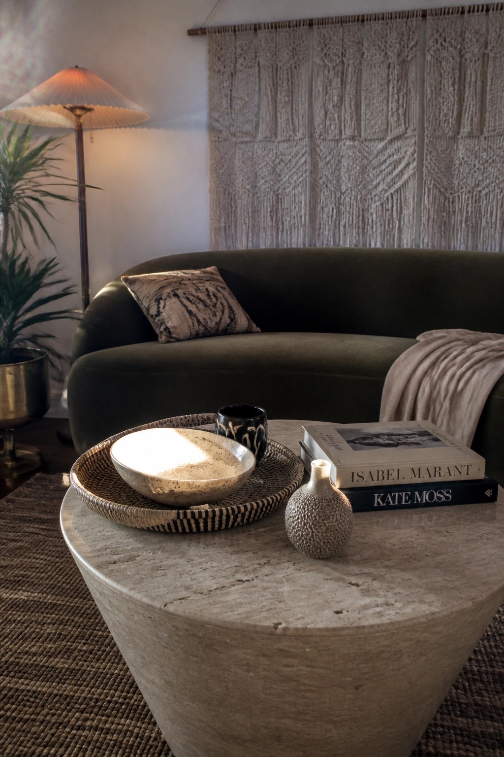 A round travertine coffee table holds a woven tray containing a speckled ceramic bowl, a small textured vase, stacked books titled Isabel Marant and Kate Moss, and a black cup, with a dark green sofa and macrame wall hanging in the background.