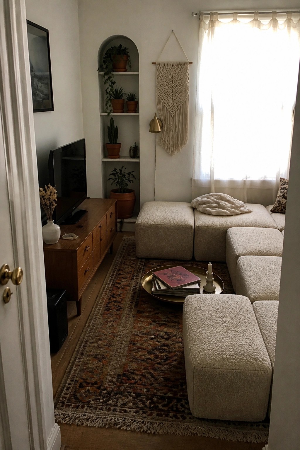 A small living room featuring white modular seating on a patterned rug, built-in arched shelves with plants, and a large window with sheer curtains.