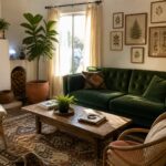 A living room with a green tufted velvet sofa, wooden coffee table on a patterned rug, two wicker chairs, and a fireplace with stacked firewood.