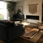 A living room with dark walls, a dark sectional sofa, round concrete coffee table, wooden cane chairs, built-in shelves, and a fireplace.