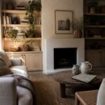 A living room corner showing built-in wooden shelves filled with books, plants, and pottery around a fireplace with a light sofa and round coffee table in front.