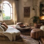 A boho living room featuring a round wooden coffee table surrounded by a rattan sofa, patterned poufs, and ottomans arranged on a large circular rug, with hanging plants, shelves, and an arched window in the background.
