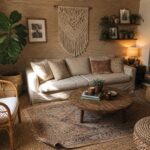 A living room with a light sofa, round wood coffee table, layered rugs including a large jute base and smaller patterned rug on top, macrame wall hanging, rattan chair, woven poufs, and multiple houseplants.