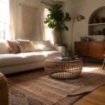 A living room with a low-profile white sofa, round rattan coffee table on a patterned rug, woven pouf, and wooden accent chair near a large window.