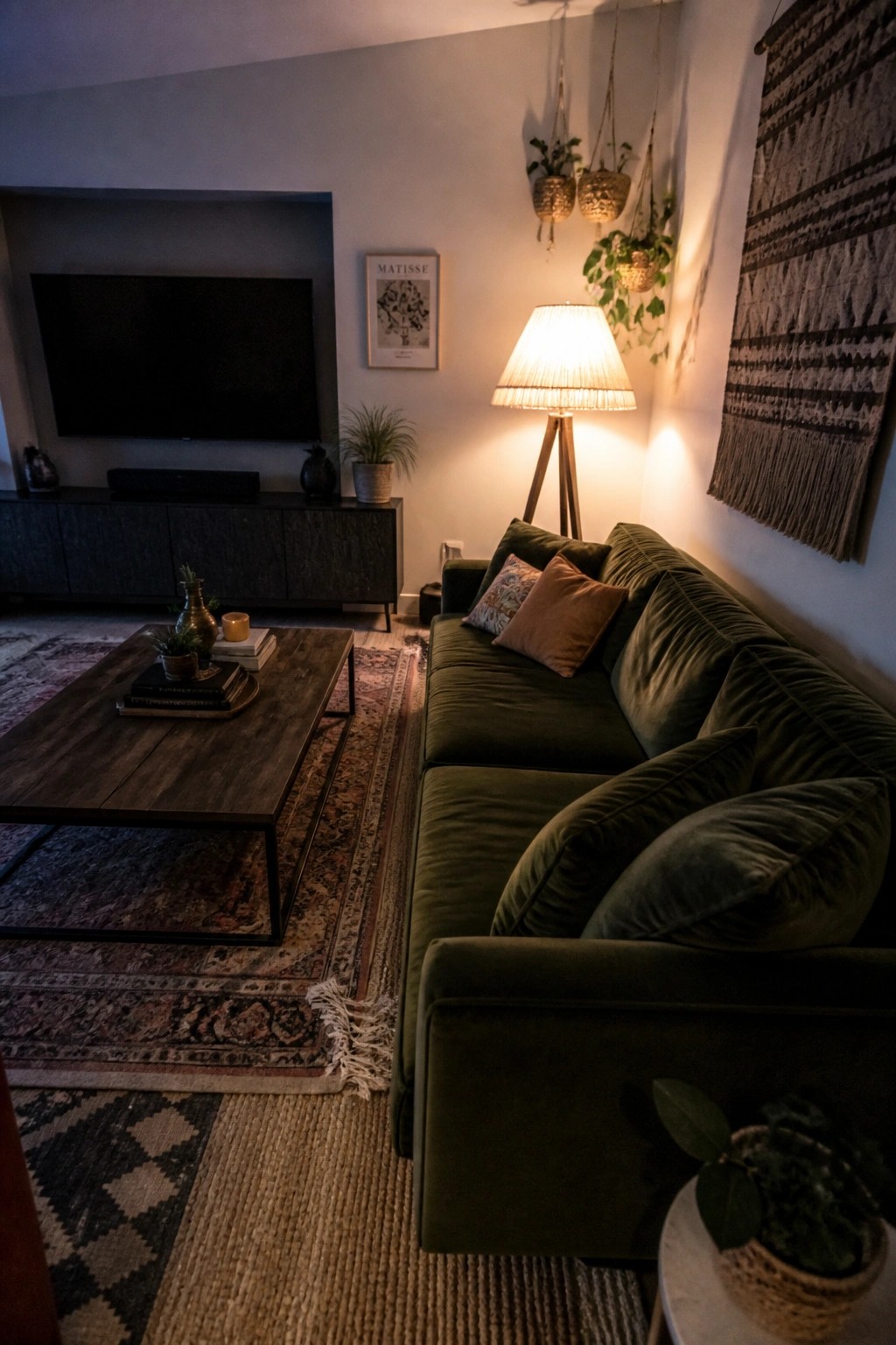 A living room featuring a dark green velvet sectional sofa, layered patterned and natural fiber rugs under a wooden coffee table, and a wall-mounted television.