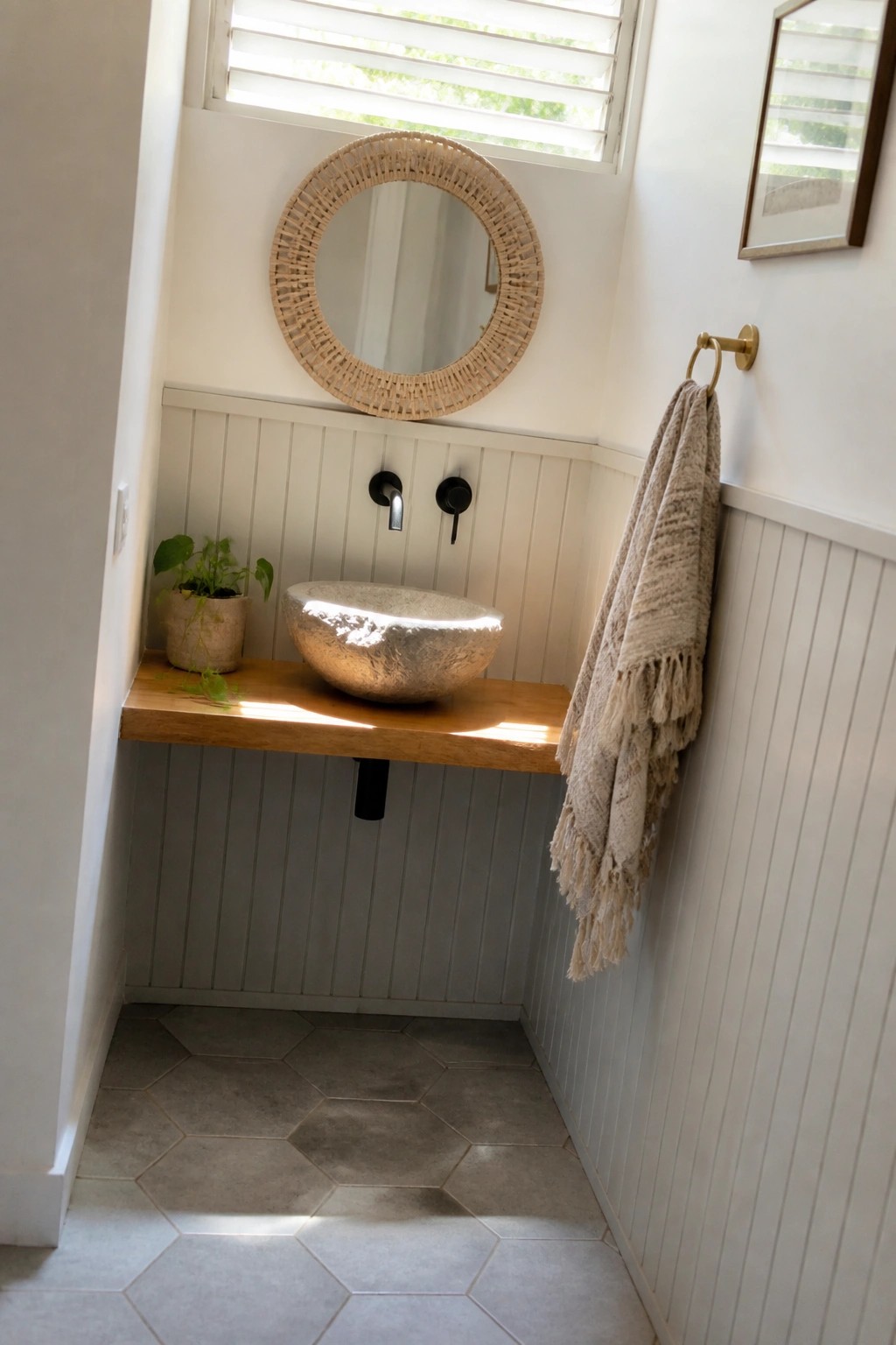 A small bathroom features a floating wooden vanity with a metallic vessel sink, black wall-mounted faucets, a round woven mirror, a potted plant on the left, and a fringed towel on a brass hook to the right.