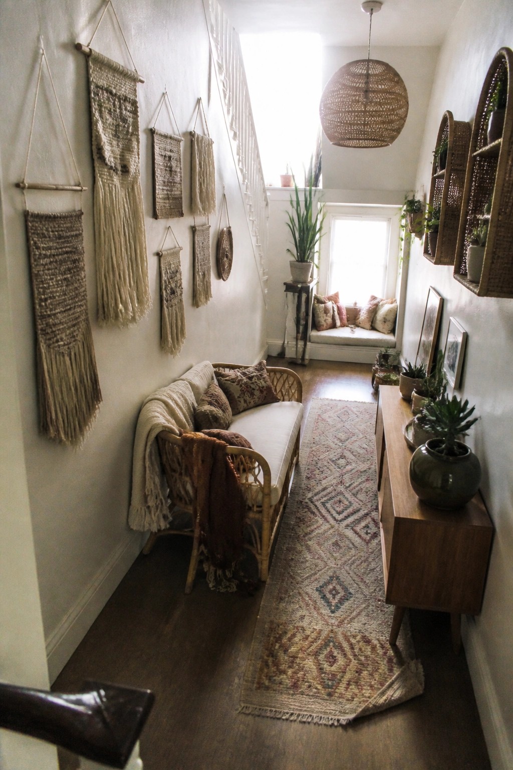 A narrow hallway interior with multiple woven macrame hangings arranged on the left wall above a rattan sofa.