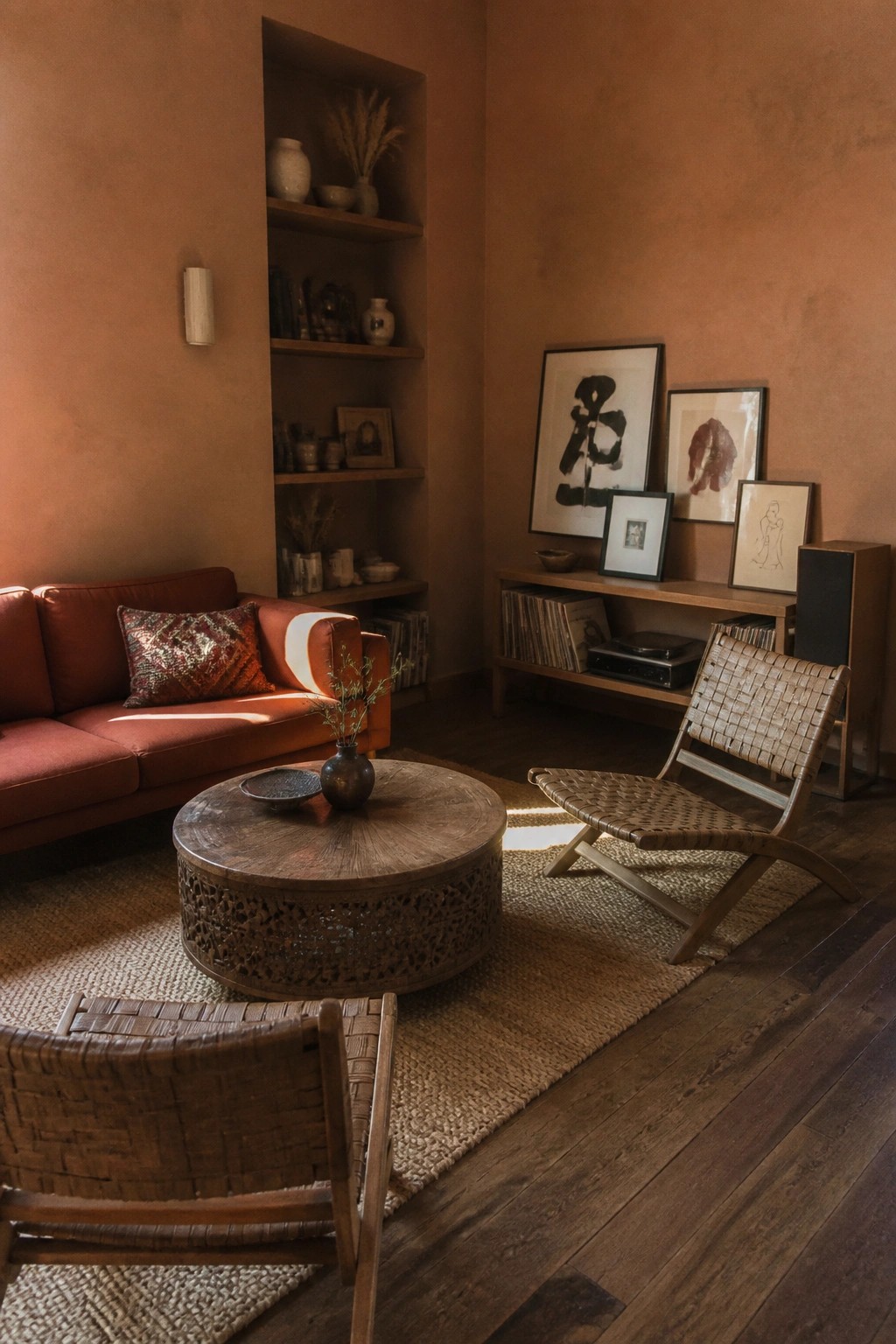 A living room with terracotta walls, a rust sofa, two woven wood chairs, a round carved coffee table, and built-in shelves filled with pottery and books.