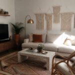 A modern boho living room with a beige sofa, patterned rug, terrazzo coffee table, and several macrame wall hangings arranged above the seating.