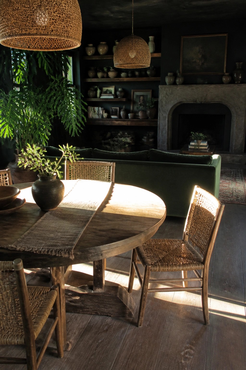 A round wooden dining table with woven chairs is placed in front of a green velvet sofa in a living room with matte black walls, open shelving filled with pottery, and a stone fireplace.