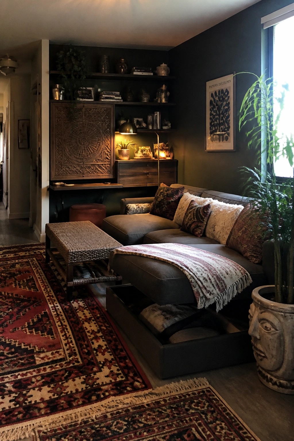 A dark gray sectional sofa with an open storage drawer under the chaise sits in a living room with dark green walls, built-in wooden shelves, patterned pillows, layered rugs, and potted plants.