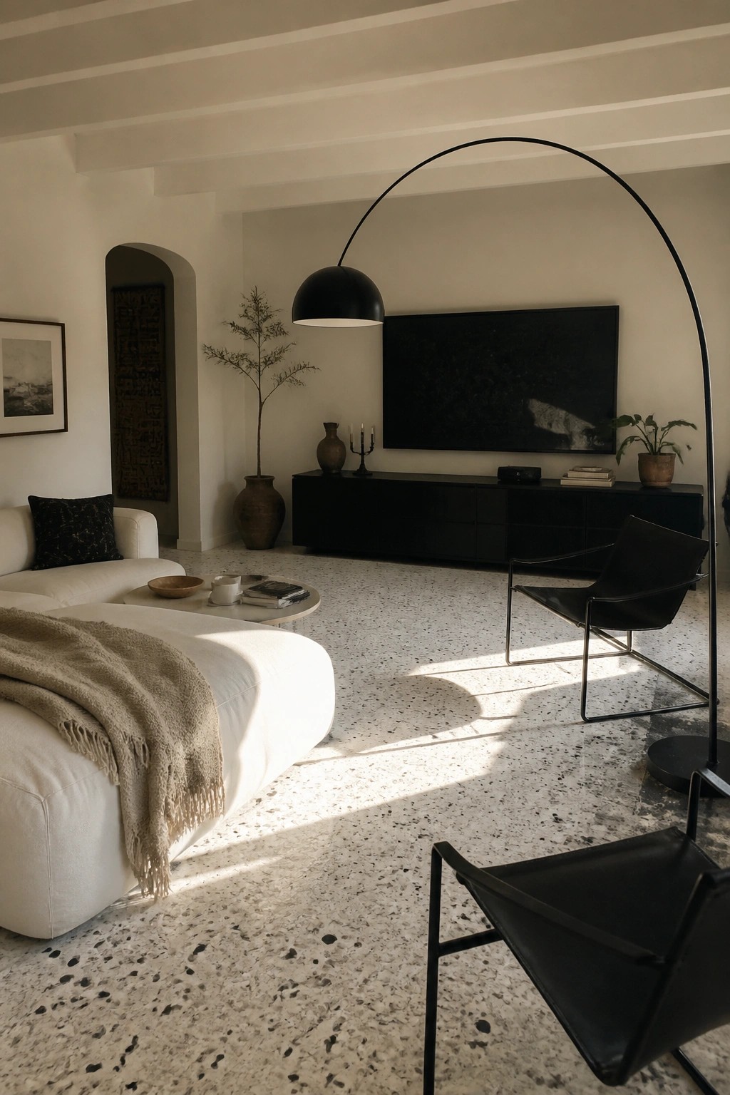 A bright modern living room featuring a white sectional sofa, two black leather chairs with metal frames, a large black arched floor lamp, and a long black media console beneath a wall-mounted television.