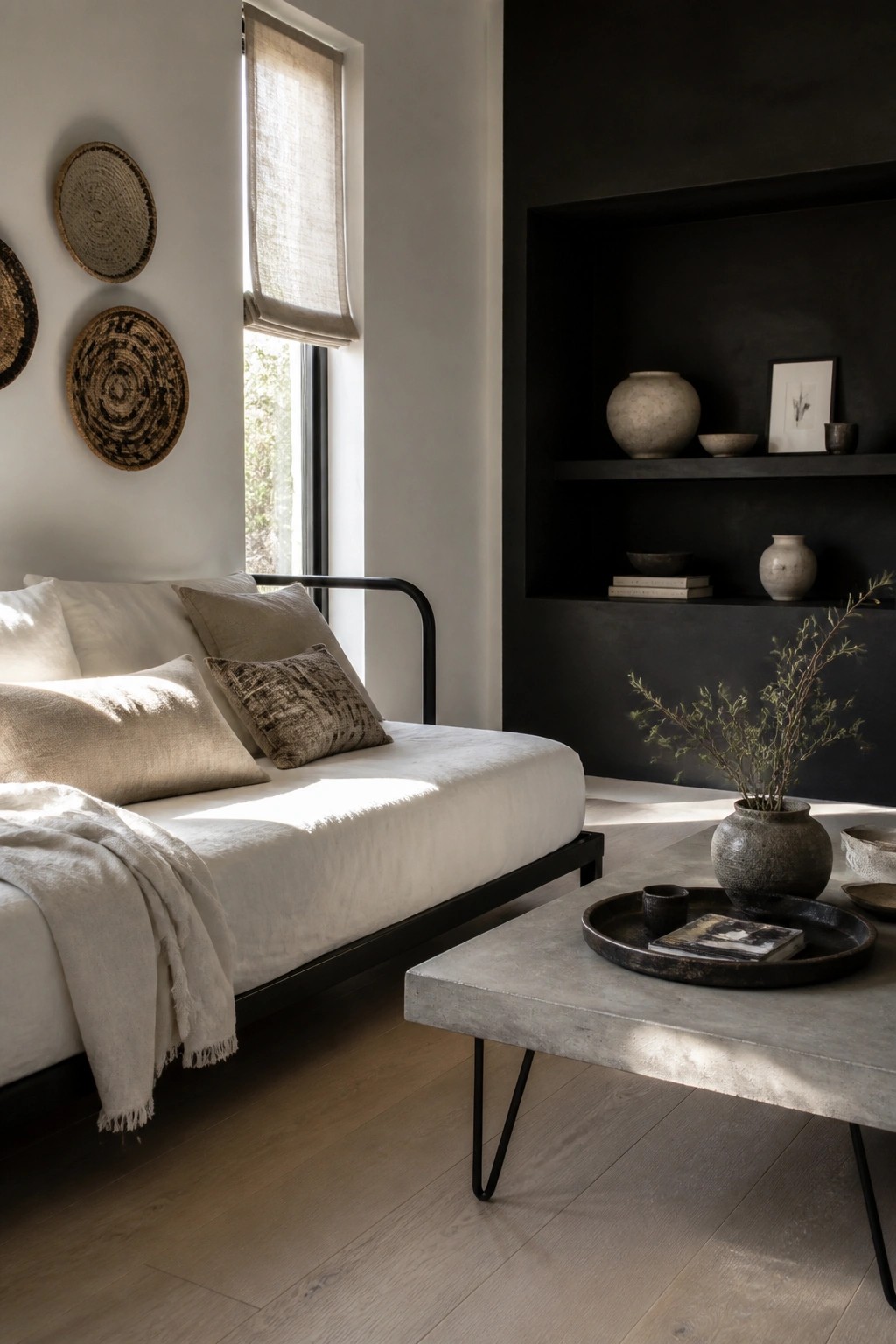 A white upholstered daybed with a black metal frame sits in a neutral room beside a concrete coffee table and dark built-in shelves holding pottery.