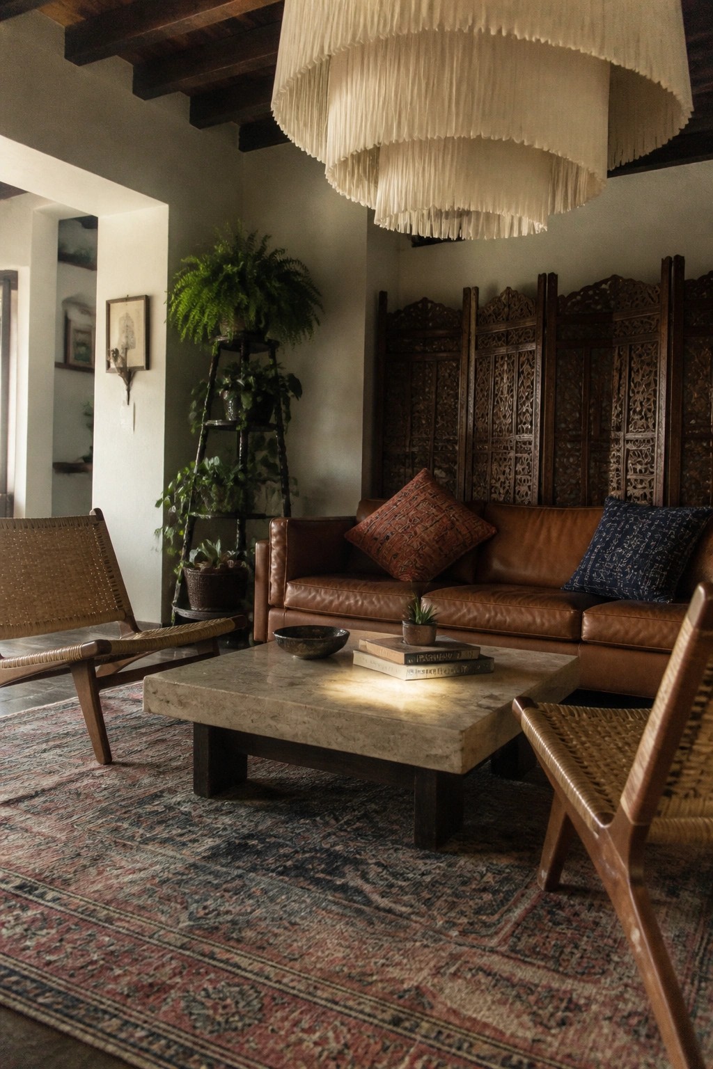 A living room with a large layered fringed pendant light hanging above a brown leather sofa, stone coffee table, patterned rug, and woven accent chairs.