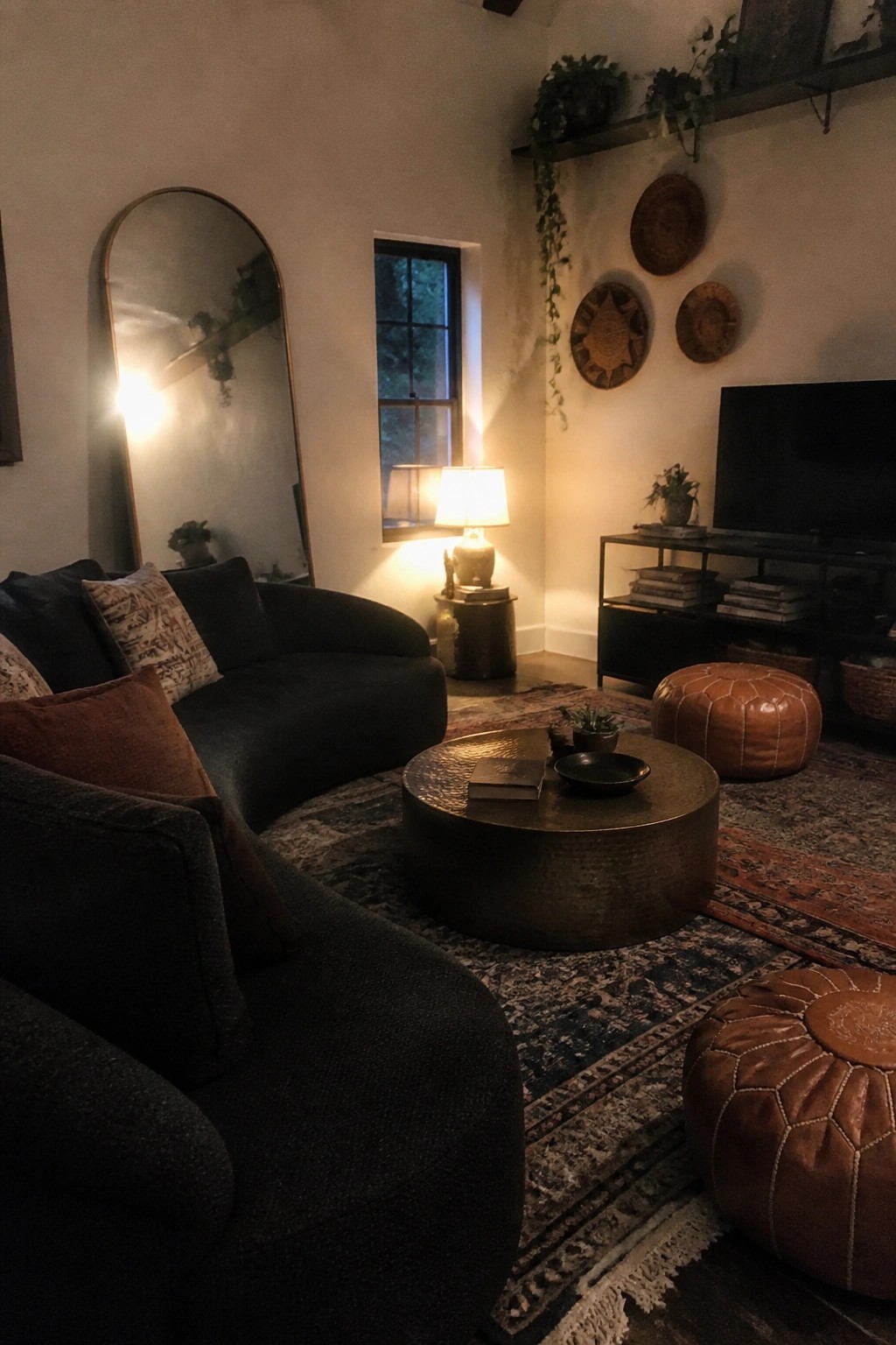 A cozy living room featuring two large dark curved sofas, a round hammered metal coffee table, leather poufs, layered rugs, and warm lamp lighting.