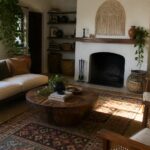 A living room featuring a white sofa, round wooden coffee table on layered rugs, fireplace with mantel, woven accent chair, and potted plants.