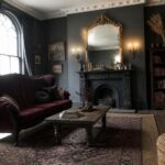 A living room featuring a red velvet sofa, dark walls, a large ornate gold mirror above a black fireplace, and a rustic wooden coffee table on a patterned rug.