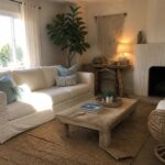 A bright living room with a white linen slipcovered sofa, wooden coffee table on a jute rug, and a wicker chair near a white fireplace.
