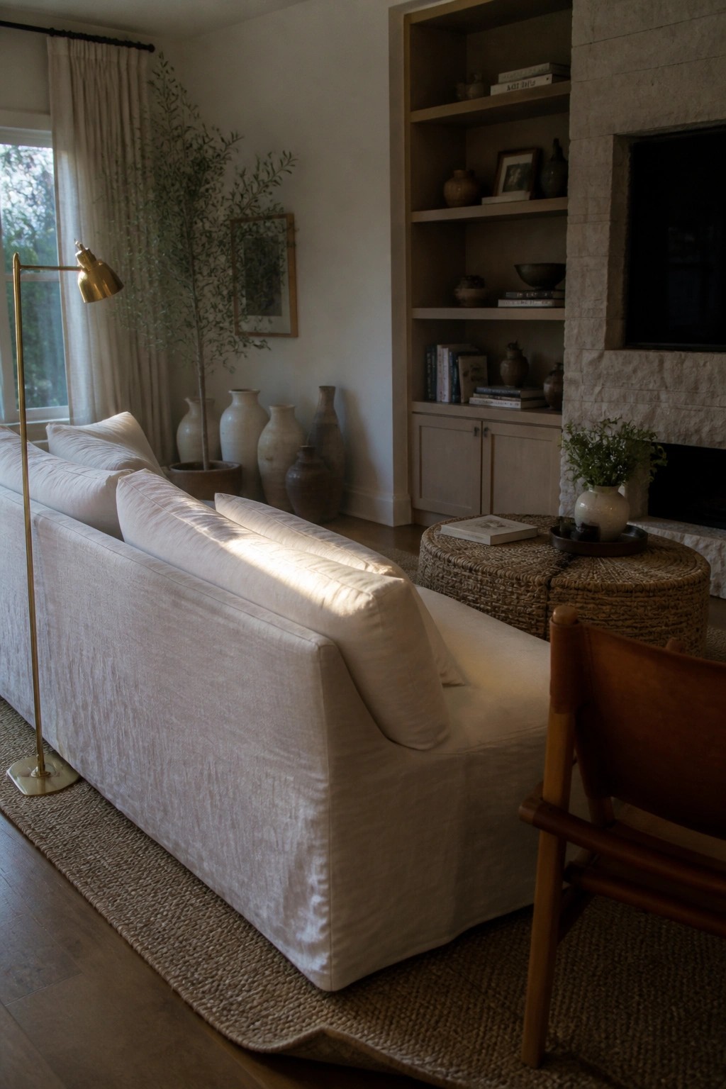 A living room with a white linen slipcovered sofa on a woven rug, a brass floor lamp beside it, built-in shelves holding books and ceramics, and a stone fireplace to the right.